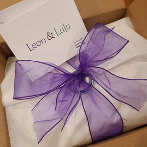 Tissue wrap[ped and tied off with wired purple ribbon into a bow. Above item is a white post card with Leon & Lulu's logo.