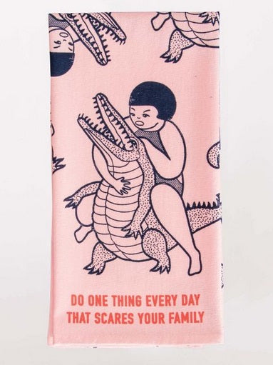 Light pink pastel screen printed dish towel. Depicted is a little girl wresting a crocodile. Below them, there is text saying, "Do one thing every day that scares your family."